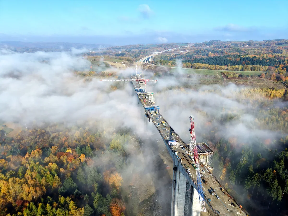 Budowa długiego mostu wiaduktu nad mglistą doliną, otoczona jesiennym lasem i polami, z dźwigami na konstrukcji.