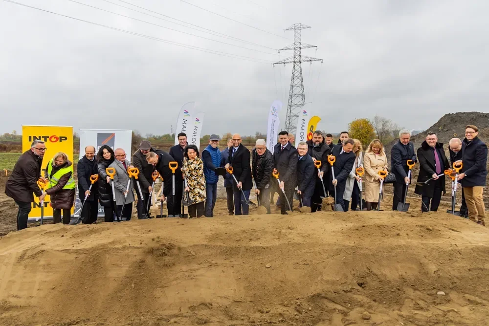 Grupa osób uczestniczy w uroczystym rozpoczęciu budowy na terenie inwestycji, wykonując symboliczne wykopy łopatami.