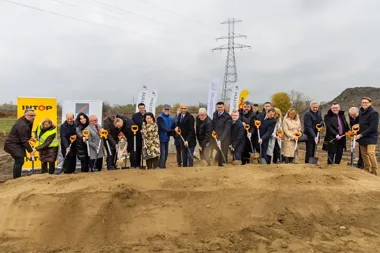 Grupa osób uczestniczy w uroczystym rozpoczęciu budowy na terenie inwestycji, wykonując symboliczne wykopy łopatami.