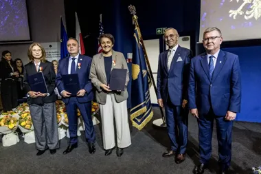 Grupa osób w strojach formalnych stoi na scenie podczas uroczystości wręczenia odznaczeń i dyplomów, w tle flagi i sztandar.