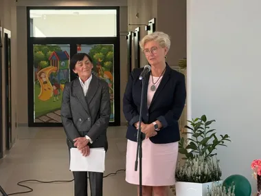 Dwie kobiety w eleganckich garniturach stoją w holu, jedna przemawia do mikrofonu, druga trzyma kartki; w tle kolorowa witrażowa ścianka z motywem placu zabaw.